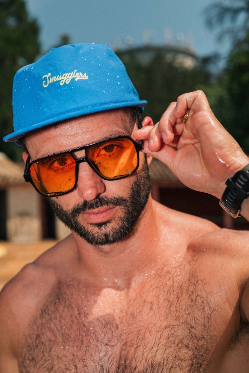 A close-up lifestyle shot of a man wearing a bright blue bucket hat with 'Swaggots' written in gold script, orange-tinted sunglasses, and a black watch. He is shirtless with water droplets on his skin, set against a blurred outdoor background.