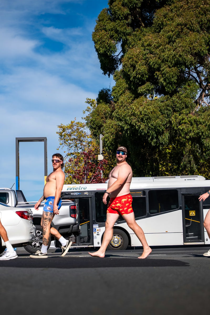 Two men walking past a city bus in underwear trunks, one in red The Hoppers kangaroo print