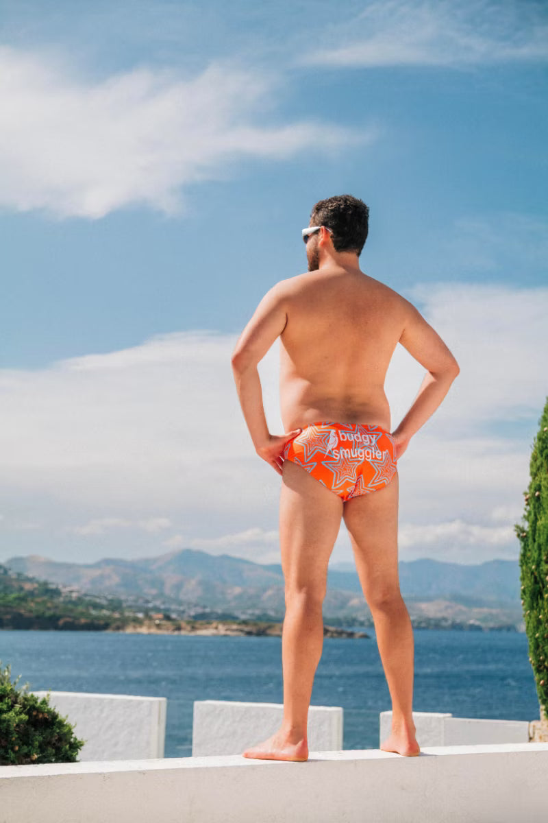 man wearing orange star swimwear brief by the water looking out. 