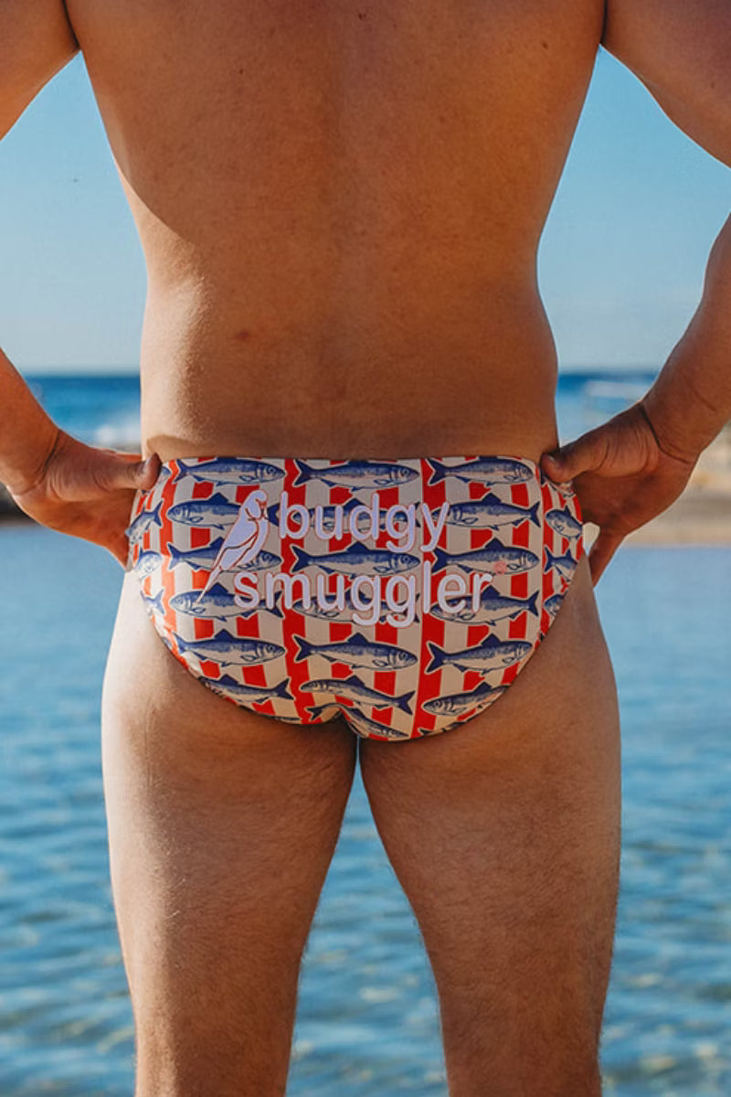 Rear view of a man standing at the beach wearing swim briefs with a repeating red, white, and blue fish pattern and a white 'Jewelry Smuggler' logo across the front.