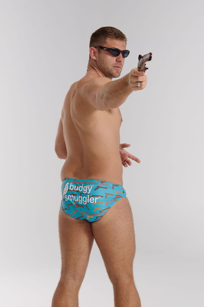Man wearing blue swimwear with 'buddy smuggler' branding, holding a stick.