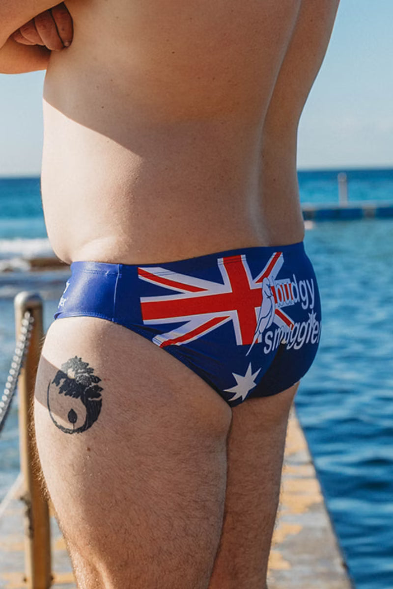 A close-up lifestyle shot of a person wearing swim briefs featuring the Australian flag and the text 'midy struggle' in white script. The person is standing on a wooden dock with the blue ocean in the background.
