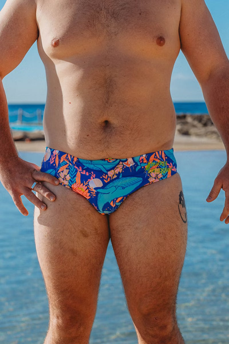 A close-up of a man wearing colorful swim briefs at the beach, with blue ocean water and sky in the background.
