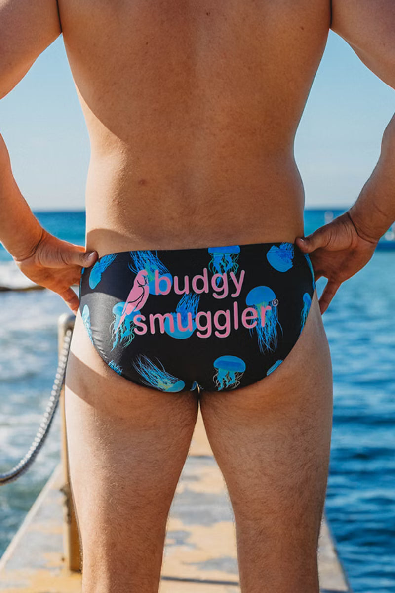 Rear view of a man wearing low-rise swim briefs with a black background, blue jellyfish pattern, and pink text reading 'budgy smuggler' on a boat with the ocean in the background.