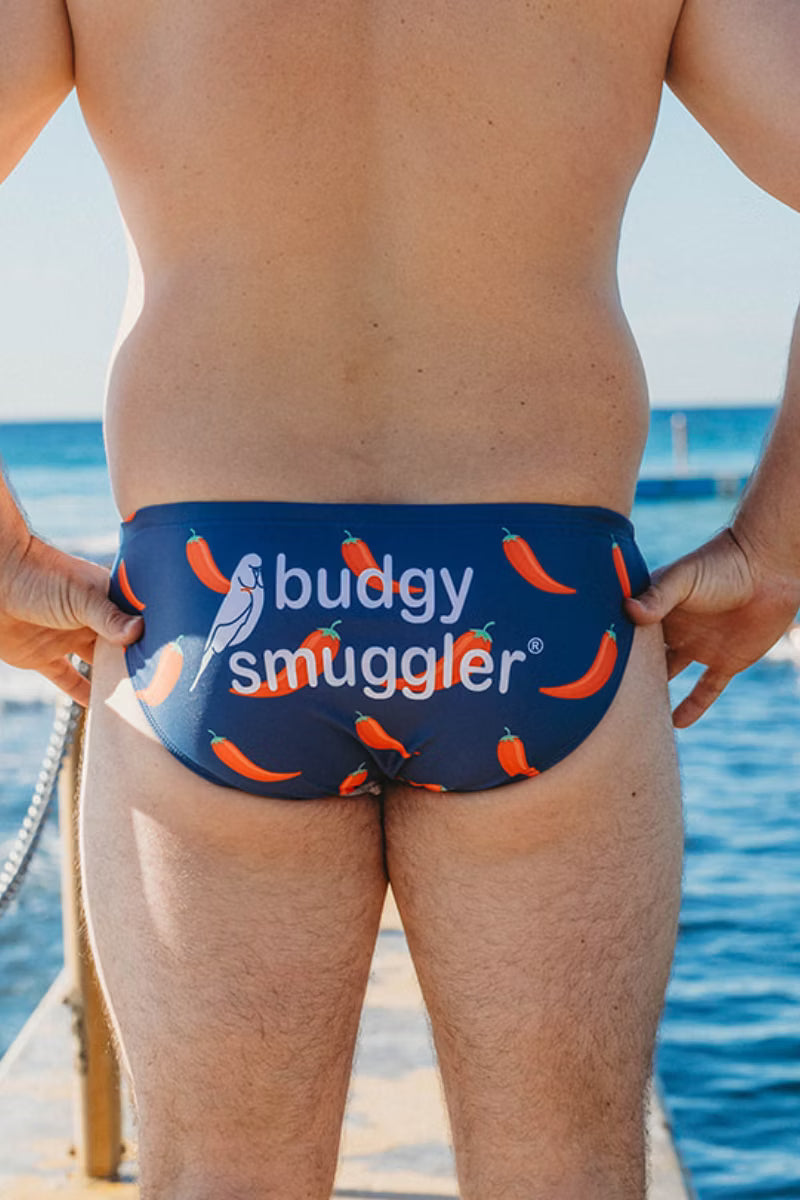 Rear view of a man wearing navy blue swim briefs with an orange chili pepper print and the white text 'budgy smuggler' on the back, standing outdoors near water.