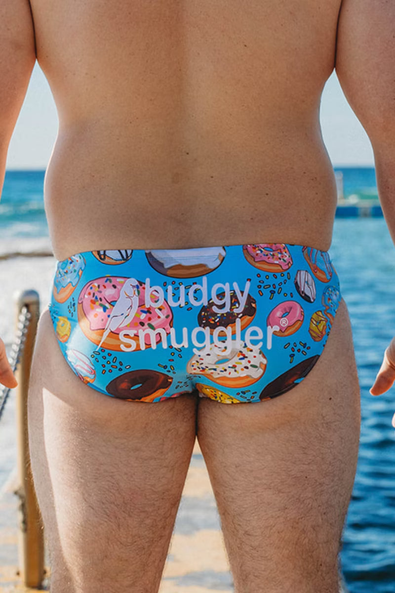 A man viewed from behind wearing bright blue swim briefs covered in a colorful donut print, standing on a beach with the ocean in the background. A semi-transparent watermark reading 'buddy smuggler' is overlaid across the center.