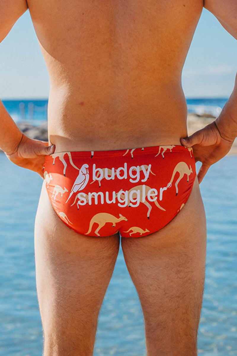 A man viewed from behind wearing red swim briefs with kangaroo and bird illustrations and the text 'buddy smuggler' on the back, standing by the ocean.