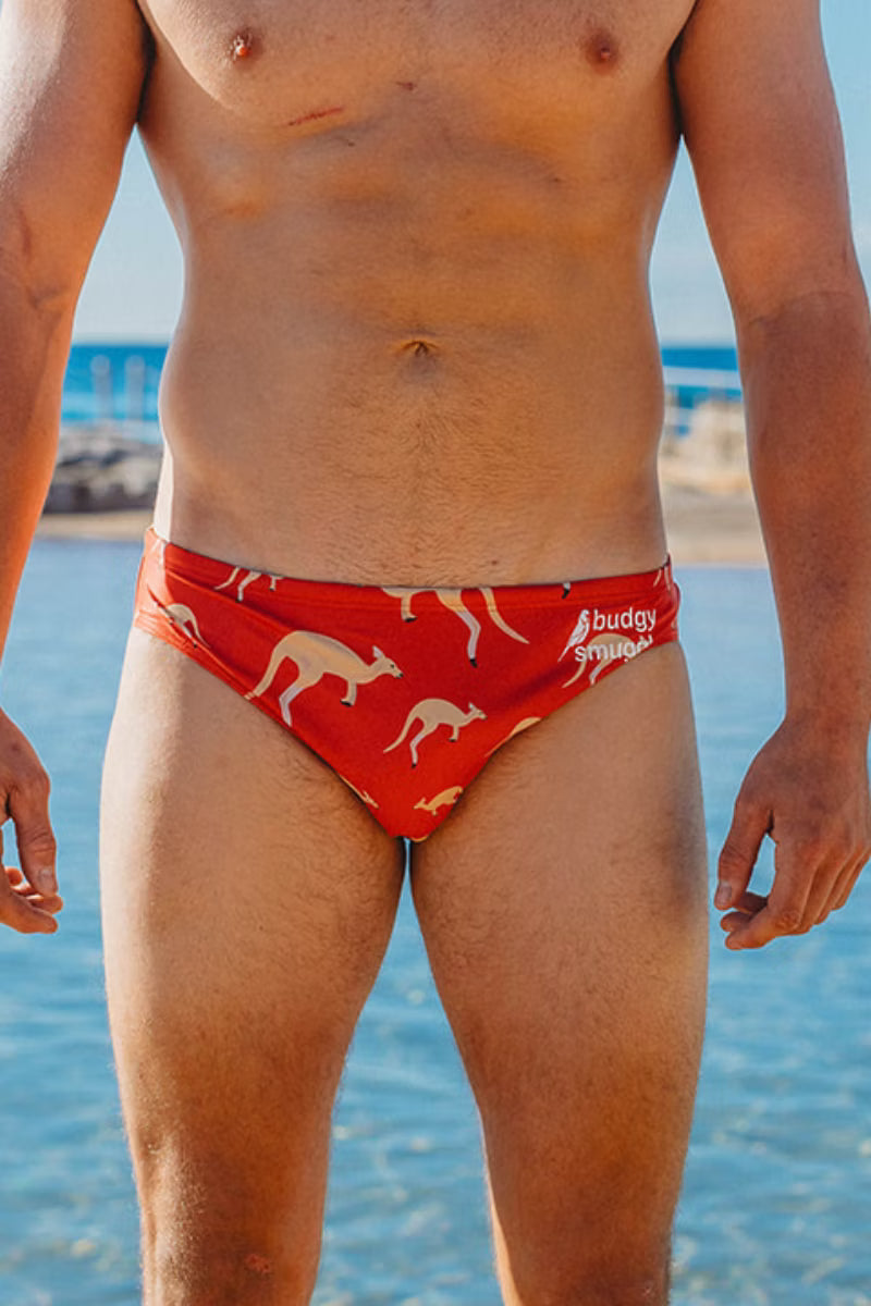 A man wearing red swim briefs with a kangaroo print, standing at the beach with water and sand in the background.