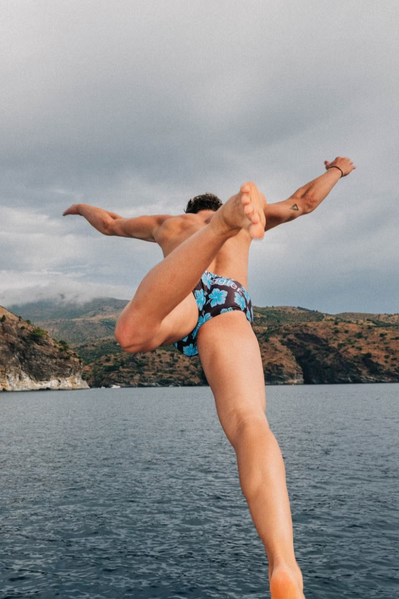 Man leaping off a boat into the sea in Brown and Blue Hibiscus swimmers, coastal cliffs behind