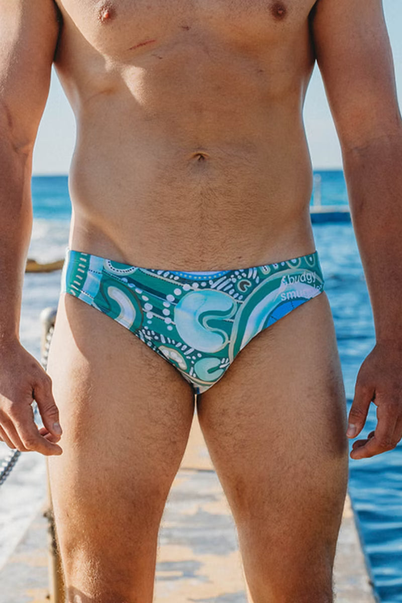 Front of the Merindah Gunya Respect swim briefs, teal and white artwork, at an ocean pool