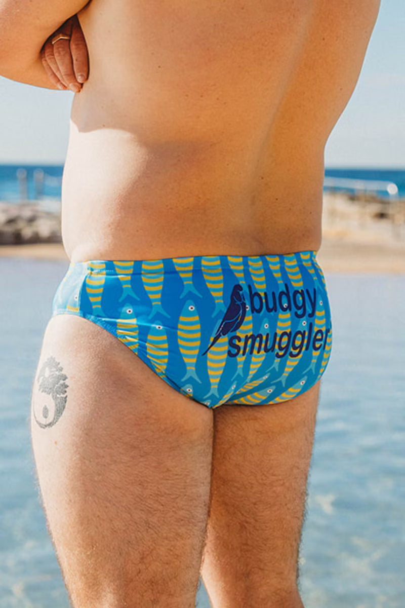A close-up back view of a person wearing blue swim briefs with a yellow fish pattern and the text 'buddy smuggler' printed on the back, standing near the beach with water in the background.
