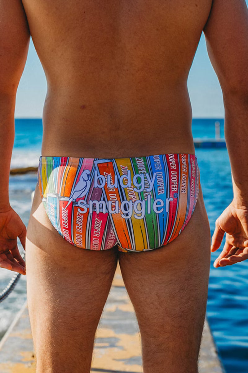 Rear view of a man wearing colorful striped swim briefs with a blue 'buddy smaggler' logo, standing outdoors near the ocean.