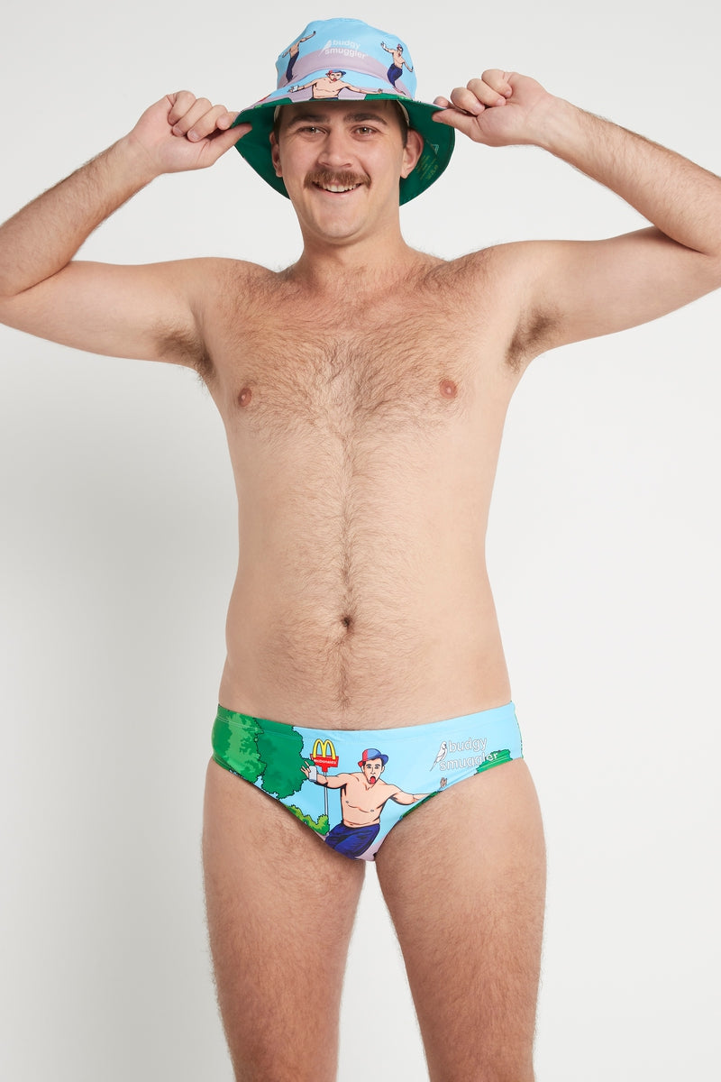 Model in a bucket hat wearing Budgy Smuggler Immortal Smugglers, Andrew Johns cartoon swim briefs, front view